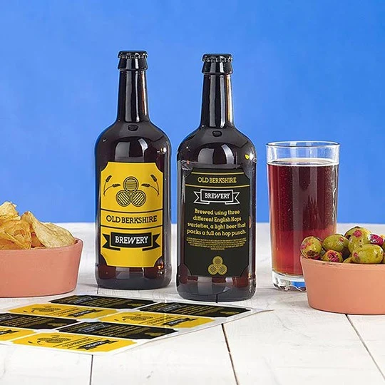 Dos botellas de cerveza con etiquetas personalizadas. Hay patatas fritas, aceitunas, vaso de cerveza y una hoja con etiquetas