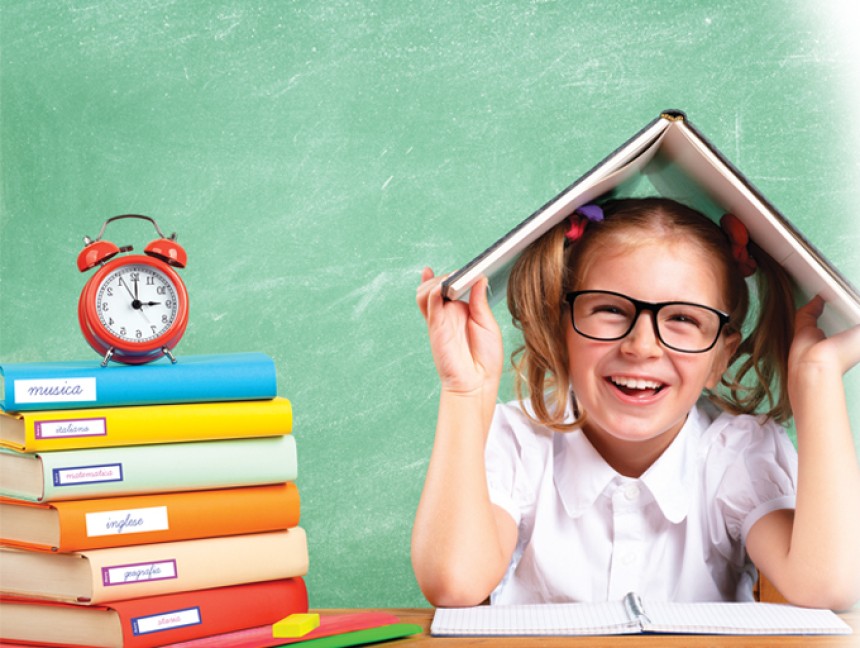 Una niña con gafas riendo con un libro sobre la cabeza y una pila de libros escolares etiquetados