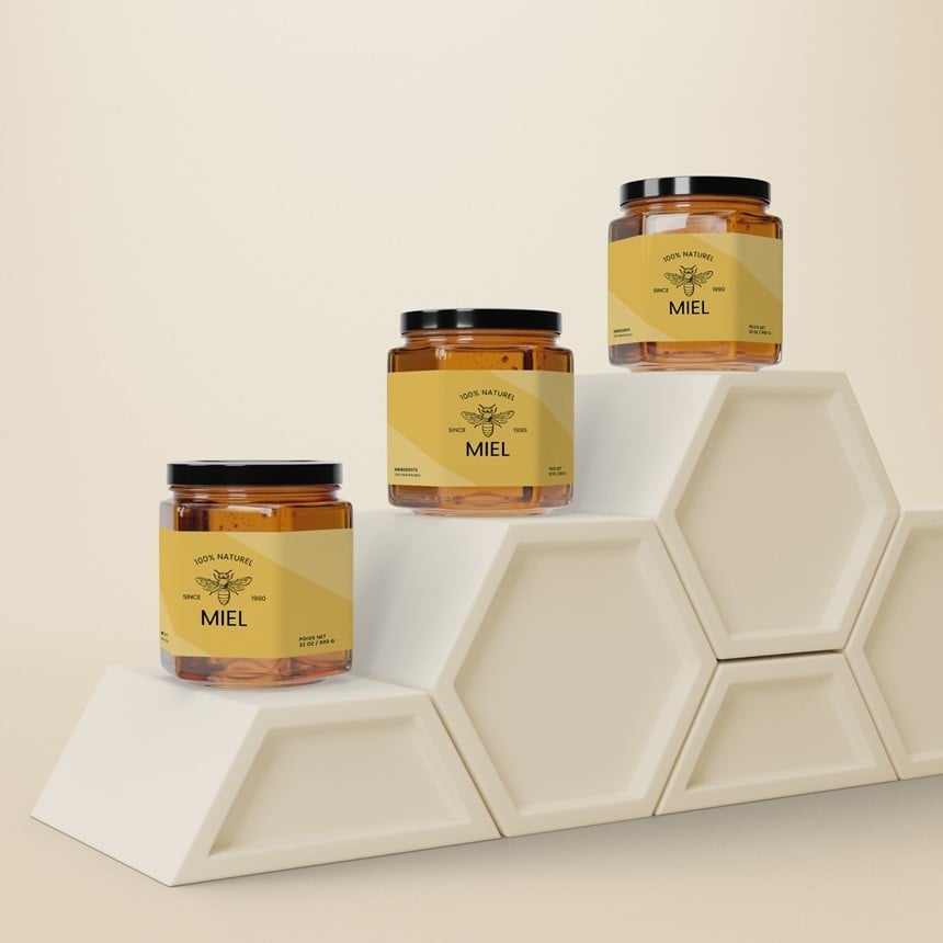 Tres tarros de miel con etiquetas envolventes rectangulares amarillas colocados en una estructura blanca con forma de panal