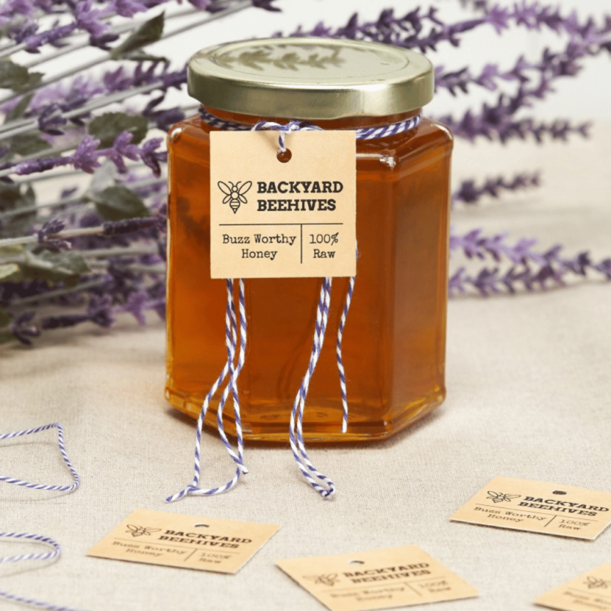 En primer plano, tarro de miel con etiqueta cuadrada colgantes apoyado sobre una mesa. Al fondo flores de lavanda