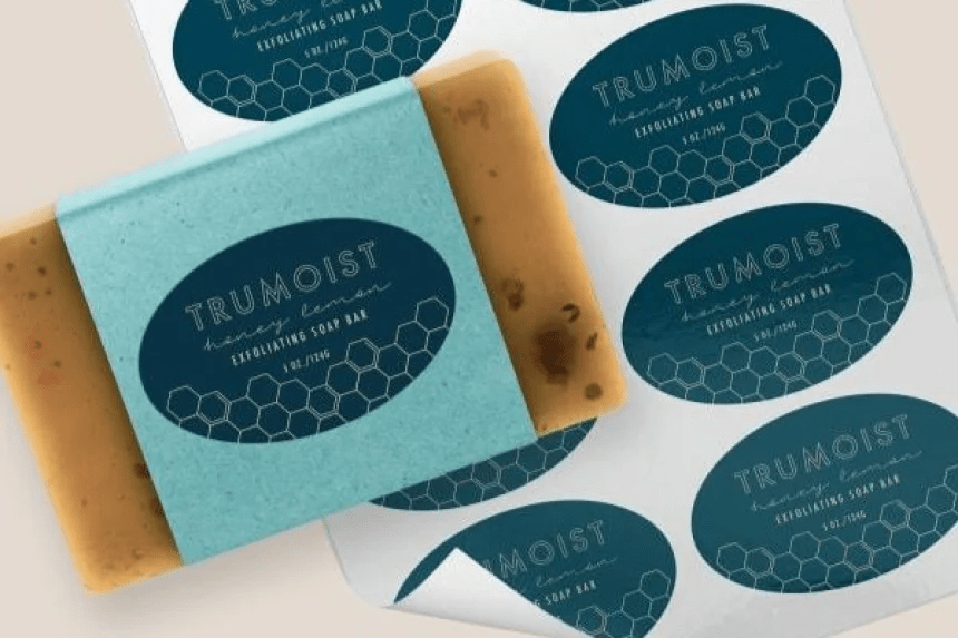 Un jabón exfoliante envuelto con una etiqueta azul ovalada. Al lado, a una hoja de etiquetas ovaladas personalizadas