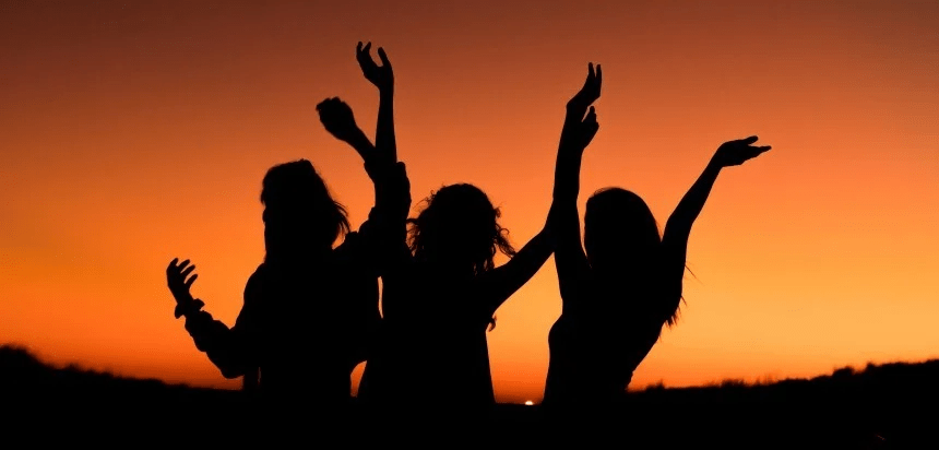 Tres siluetas de mujeres con los brazos levantados contra una puesta de sol naranja y amarilla