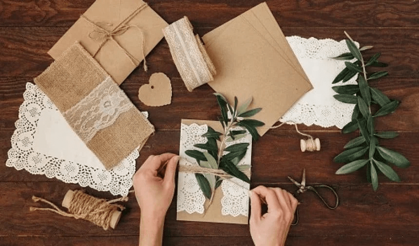 Manos creando invitación de boda rústica con papel kraft, encaje, cuerda, corazón de cartón y ramas de olivo sobre mesa de madera