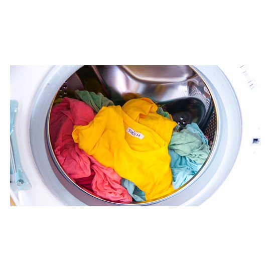La imagen muestra una lavadora llena de ropa de varios colores, incluyendo una camiseta amarilla y otras prendas en tonos rosa y azul claro, girando dentro del tambor.