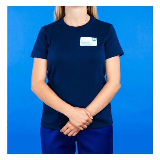 La imagen muestra a una mujer de pie con una camiseta azul marino y pantalones, frente a un fondo azul. Lleva una etiqueta con su nombre y cargo, sosteniendo las manos en una posición neutra.