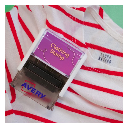 La imagen muestra un sello para ropa de la marca Avery, colocado sobre una camiseta de rayas rojas y blancas. El sello está diseñado para marcar prendas con el texto 'Clothing Stamp'.