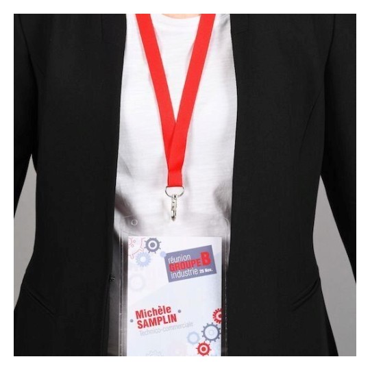 La imagen muestra a una persona con un abrigo negro y una camiseta blanca, usando un gafete con un cordón rojo. El gafete tiene detalles en azul y rojo y parece estar relacionado con un evento industrial.