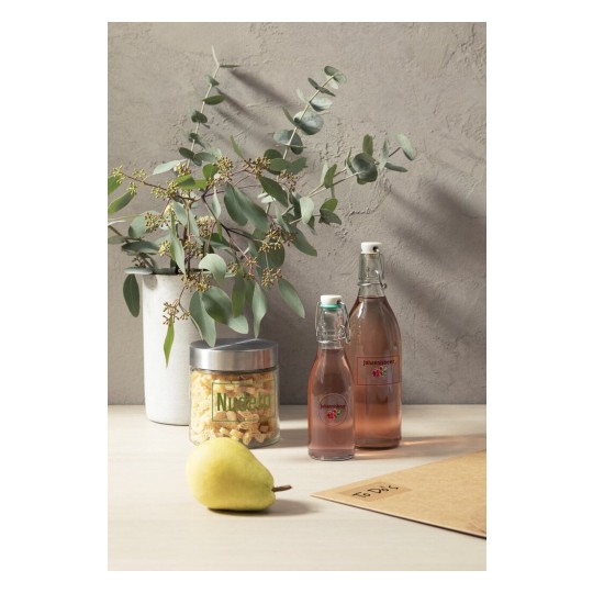 La imagen muestra una composición con una planta de eucalipto, dos botellas con líquido, un frasco con pasta, una pera y un sobre en una mesa.