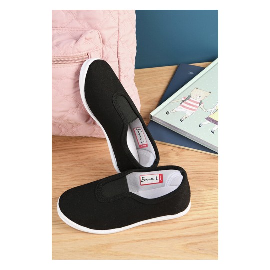 La imagen muestra un par de zapatos negros de tela con suela blanca, colocados sobre una superficie de madera. Al fondo se ve una mochila rosa y libros infantiles con ilustraciones.