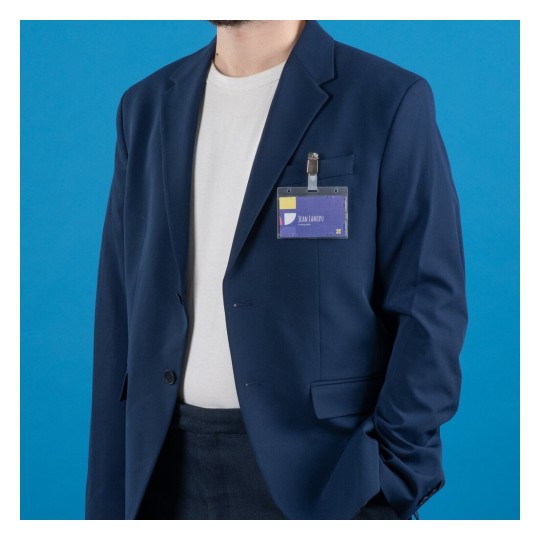 Una persona con un traje azul oscuro y una camiseta blanca, llevando un gafete con su nombre. El fondo es azul.