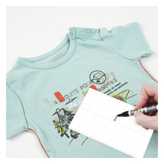 La imagen muestra una camiseta azul claro con un diseño gráfico y una etiqueta blanca donde se está escribiendo un nombre. La camiseta parece ser de niño o bebé.