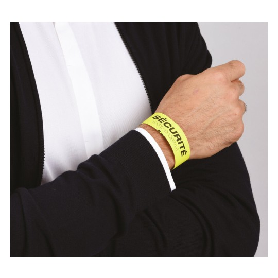 En la imagen se observa una mano que lleva una pulsera amarilla con la palabra 'SÉCURITÉ'. La pulsera parece ser de un evento o actividad deportiva, y la persona está vestida con un suéter oscuro sobre una camisa blanca.