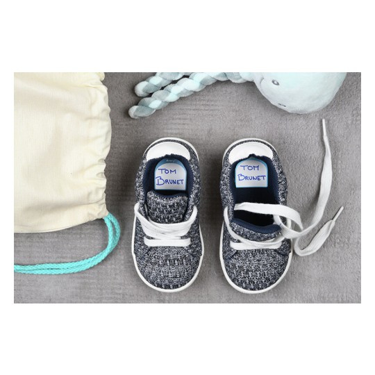 La imagen muestra un par de zapatitos pequeños de color gris oscuro con cordones blancos, un saco de tela claro y un juguete suave de color azul. Los zapatitos tienen el nombre 'TOM BRUNET' escrito en sus etiquetas.