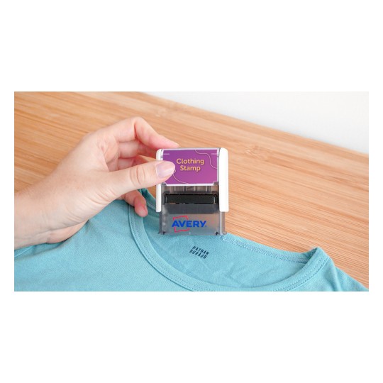 Una persona está utilizando un sello de ropa sobre una camiseta azul, que está sobre una superficie de madera clara. El sello dice 'Clothing Stamp' y es de la marca Avery.