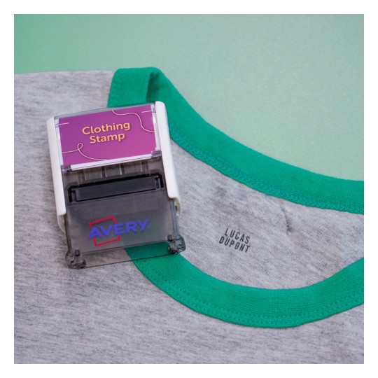 La imagen muestra un estampador de ropa con la etiqueta 'Clothing Stamp' en una camiseta gris con cuello verde. También se puede ver el nombre 'LUCAS DUPONT' impreso en la camiseta.