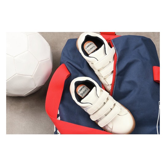 En la imagen se pueden ver un balón de fútbol, un par de zapatillas deportivas blancas y una bolsa de deporte azul con rojo. Las zapatillas tienen etiquetas visibles con el nombre 'Gabriel' y 'Judo'.