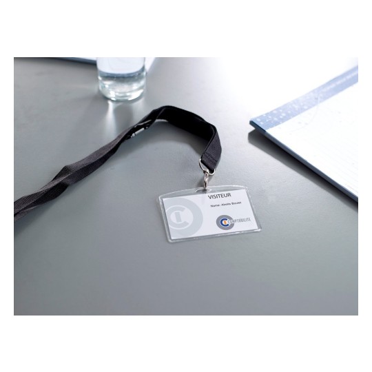 En la imagen hay una etiqueta de visita con un cordón negro, que muestra la palabra 'VISITEUR' y el nombre 'Abella Boulet' sobre una mesa. También hay un vaso de agua y un bloc de notas al fondo.