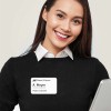 Mujer sonriendo con suéter negro y camisa blanca, lleva un identificativo adhesivo  con su nombre