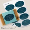Un jabón exfoliante envuelto con una etiqueta azul ovalada.  Al lado, a una hoja de etiquetas ovaladas personalizadas