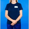 La imagen muestra a una mujer de pie con una camiseta azul marino y pantalones, frente a un fondo azul. Lleva una etiqueta con su nombre y cargo, sosteniendo las manos en una posición neutra.