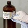 La imagen muestra una botella de solución de cloruro férrico con una etiqueta que contiene advertencias y precauciones. Una mano con un guante blanco está limpiando el frasco con un paño azul.