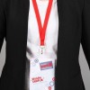 La imagen muestra a una persona con un abrigo negro y una camiseta blanca, usando un gafete con un cordón rojo. El gafete tiene detalles en azul y rojo y parece estar relacionado con un evento industrial.