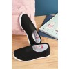 La imagen muestra un par de zapatos negros de tela con suela blanca, colocados sobre una superficie de madera. Al fondo se ve una mochila rosa y libros infantiles con ilustraciones.