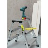 La imagen muestra una escalera de aluminio con un destornillador colocado en ella. También hay una regla amarilla y algunas tablas de madera en el fondo.