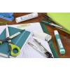 Una imagen que muestra varios útiles escolares desordenados sobre una mesa, incluyendo tijeras, una libreta, un lápiz, pegamento, un borrador, un compás y un bolígrafo en un estuche verde.