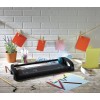 La imagen muestra una mesa con herramientas de manualidades, incluyendo una cortadora de papel, hojas de colores y una planta en maceta. Se ven tarjetas colgadas con pinzas en una cuerda.