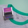 La imagen muestra un estampador de ropa con la etiqueta 'Clothing Stamp' en una camiseta gris con cuello verde. También se puede ver el nombre 'LUCAS DUPONT' impreso en la camiseta.
