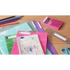 La imagen muestra varios cuadernos, una calculadora rosa, una regla, tijeras y lápices de colores sobre una mesa de madera.