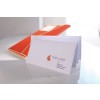 En la imagen se puede ver un sobre blanco con una tarjeta de presentación y un archivo de color naranja. El sobre lleva información de contacto sobre una firma de asesoría y está colocado sobre una superficie brillante.